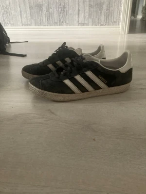 Adidas Gazelle svarta sneakers - Klassiska Adidas Gazelle sneakers i svart mocka med vita detaljer och de ikoniska tre ränderna på sidan. Låg modell med vit sula och guldfärgad Gazelle-text på sidan. Perfekta för en stilren och sportig look.
