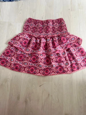 Rosa kjol - Kort kjol med rosa och röda blommor, bred smock i midjan och volangdetalj. Perfekt för dig som gillar färgglada och lekfulla plagg. Kjolen har en luftig passform och är gjord i ett mjukt bomullsliknande material.