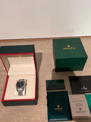 Role klocka med svart urtavla - Säljer en stilren Role klocka i silver med svart urtavla och länkarmband. Klockan har en bezel med stenar och tydliga indexmarkeringar. Kommer med originalbox, certifikat och tillbehör. Perfekt för dig som vill ha en klassisk och lyxig accessoar.