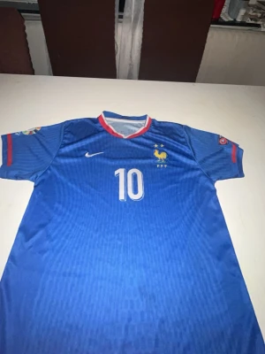 Frankrike fotbollströja Mbappé #10 - Blå fotbollströja från Nike med Frankrikes landslagsmärke och nummer 10 på bröstet. På ryggen står det 'Mbappé' och siffran 10. Tröjan har röda och vita detaljer vid kragen och ärmarna samt tryck på båda ärmarna. Perfekt för dig som älskar fotboll och vill stötta Frankrike.