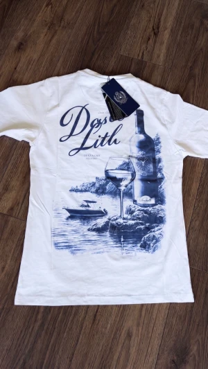 Marcél T-Shirt Vit  -  Vit t-shirt från  Marcél med stort blått tryck på ryggen vinflaska, glas och havsmotiv med texten Dolce Lith. Liten logga på bröstet fram. Klassisk passform, sitter snyggt till jeans eller shorts.