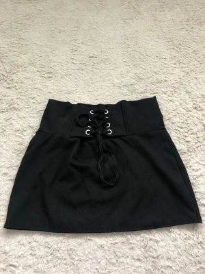 Svart kortkjol med snörning i midjan - Säljer en svart kortkjol med bred midja och snygg snörningsdetalj framtill. Kjolen har en enkel, stilren design och passar perfekt till en trendig vardagslook. Materialet känns mjukt och stretchigt för skön passform.
