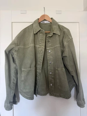 Olivgrön jeansjacka från Zara - Olivgrön jeansjacka från Zara med avslappnad passform, stora framfickor och klassisk krage. Jackan har silverfärgade knappar och långa ärmar med knappdetalj vid ärmslut. Perfekt för en cool och avslappnad stil.