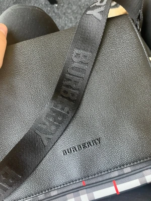 Svart axelväska från Burberry - Snygg svart axelväska från Burberry i strukturerat läder med justerbar axelrem där Burberry-loggan är invävd. Väskan har klassiskt rutigt mönster i beige, svart, vitt och rött på baksidan samt magnetknappar. Perfekt för dig som gillar stilrena detaljer och ikonisk design.