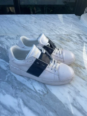 Valentino Garavani - Snygga vita sneakers från Valentino Garavani med bred svart rem över snörningen och guldfärgad logga på plösen. Skorna har klassisk låg profil och är tillverkade i läder med detaljerade nitar på hälen. Perfekta för dig som gillar exklusiv streetstyle.