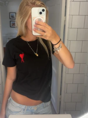Svart t-shirt från Ami Paris - Säljer en svart t-shirt från Ami Paris med rött hjärta och bokstaven A broderat på bröstet. Klassisk passform med korta ärmar och rund hals. Perfekt basic med en snygg detalj framtill. Ej äkta