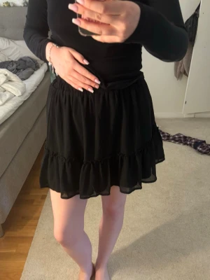 Svart volangkjol med resårmidja - Snygg svart kortkjol med volangdetaljer och resår i midjan. Kjolen har en luftig och flowy känsla, perfekt för att styla med en enkel topp. Materialet känns lätt och bekvämt.