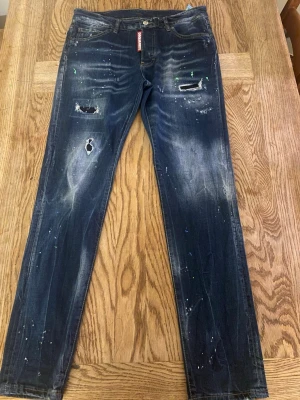 Mörkblå distressed jeans Dsquared2 - Säljer ett par mörkblå jeans från Dsquared2 med slitningar, hål och färgstänk för en riktigt edgy look. Smal passform och klassisk femficksmodell. Perfekta för dig som gillar streetstyle och vill sticka ut lite extra.