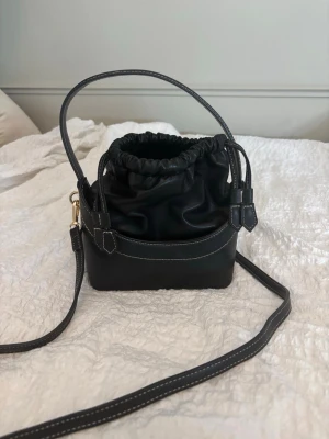 Svart bucket bag från H&M - Trendig svart bucket bag från H&M med justerbar axelrem och handtag. Väskan har snygga kontrastsömmar och dragsko upptill som ger en cool look. Perfekt för dig som vill ha en stilren och praktisk handväska i syntetmaterial.