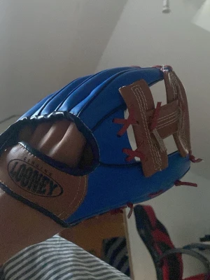 Blå basebollhandske Looney - Säljer en blå basebollhandske från Looney med bruna detaljer och röda snören. Handsken har en tecknad basebollfigur på insidan och är tillverkad i konstläder. Perfekt för dig som gillar sport och vill ha en färgglad handske.