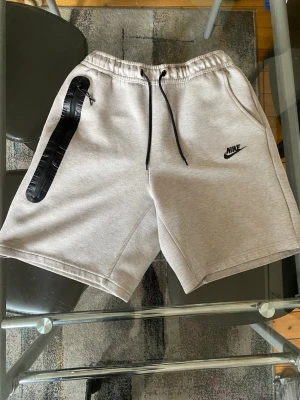Nike shorts - Snygga ljusgrå sweatshorts från Nike med svart logga på vänster ben och svarta dragsnören i midjan. Shortsen har en relaxed passform, elastisk midja och praktiska fickor. Perfekta för chill dagar eller träning.