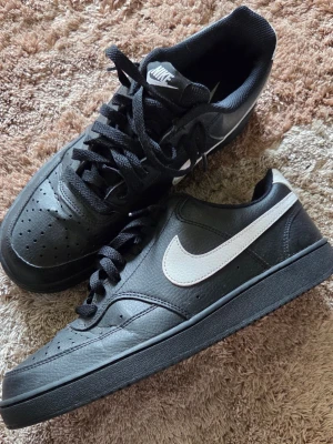 Svarta Nike sneakers med vit swoosh - Snygga svarta Nike Court vision. Skorna har klassisk snörning, perforerad tå och slitstark yttersula med cirkulärt mönster. Tillverkade i läderliknande material och textilfoder. Perfekta för en streetwear-look.