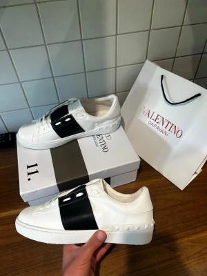 Valentino Garavani vita sneakers - Snygga vita sneakers från Valentino Garavani med bred svart läderdetalj över mitten och nitar längs hälen. Skorna har klassisk låg profil, vita snören och en robust gummisula med mönstrad undersida. Perfekta för dig som gillar exklusiv och stilren design.Väldigt fin och snygg för sommaren och för träningen. Fraktar så fort du har köpt🚚📦Aktiv 24/7✅ Hör av dig via frågor⁉️