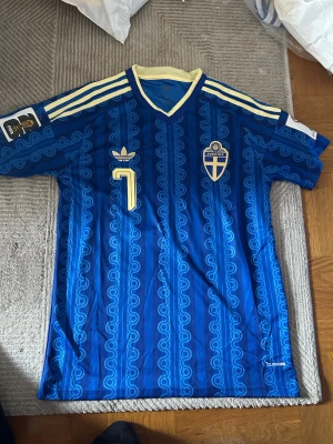 Blå Sverige retro fotbollströja Adidas - Retro fotbollströja från Adidas i blått med gula detaljer och nummer 7 på bröstet. Tröjan har Sveriges landslagsemblem, klassiska Adidas-ränder på axlarna och ett unikt mönster i tyget. Tillverkad i lätt och ventilerande material, perfekt för match eller samling.