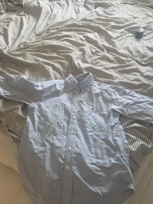 Ljusblå skjorta från Polo Ralph Lauren - Klassisk ljusblå skjorta från Polo ralphlauren passar till 12 åriga. Skjortan har knappar hela vägen och button-down krage. Perfekt till jeans eller chinos för en clean och stilren look.