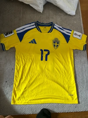 Svensk landslagströja Adidas gul - Gul svensk landslagströja från Adidas med blå detaljer och nummer 17 på bröstet. Tröjan har Sveriges landslagsemblem, blåa ränder på axlarna och vit/blå krage. Tillverkad i lätt och ventilerande material, perfekt för fotboll eller träning.