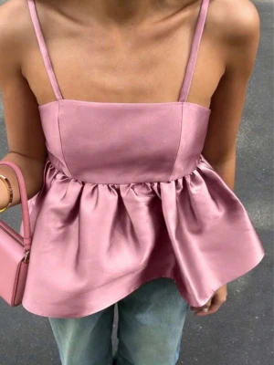 Rosa satinlinnetopp med volang - Supersöt rosa linnetopp i glansigt satinmaterial med tunna axelband och volang nedtill. Toppen har en croppad passform och ger en riktigt trendig vibe. Perfekt att styla med jeans eller kjol för en cool look.