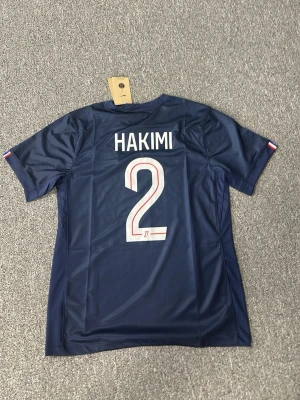 PSG matchtröja Hakimi #2 Nike - Snygg mörkblå PSG matchtröja med Hakimi och nummer 2 på ryggen. Tröjan har korta ärmar, röd och vit rand med mönster framtill, samt PSG-logga och Nike-logga på bröstet. Tillverkad i lätt och ventilerande polyester, perfekt för fotboll eller träning.