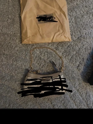 Beige miniväska med guldkedja  - Snygg beige minihandväska  med klassiskt monogrammönster och guldfärgade detaljer. Väskan har en kedjerem i guld och dragkedja upptill. Perfekt för det viktigaste och ger en lyxig touch till din outfit.