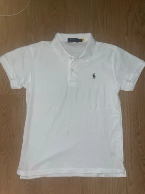 Vit pikétröja från Polo Ralph Lauren - Klassisk vit pikétröja från Polo Ralph Lauren med korta ärmar och den ikoniska broderade loggan på bröstet. Tröjan har krage och knappar framtill. Perfekt till jeans eller shorts för en clean och stilren look.