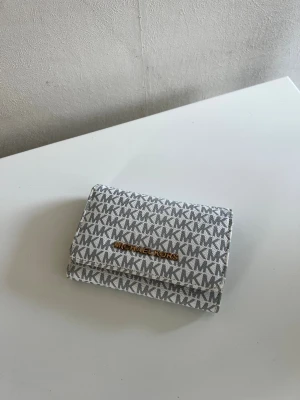 Grå Michael Kors plånbok - Snygg grå plånbok från Michael Kors (ej äkta) med MK-logga över hela ytan och guldfärgad text framtill. Plånboken har flera kortfack, myntfack med dragkedja och praktisk knappstängning. Tillverkad i tåligt konstläder med beige insida.