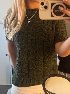 Mörkgrön kabelstickad topp - Snygg mörkgrön kabelstickad topp med korta ärmar och rund hals. Tröjan har ett klassiskt flätmönster framtill och ribbade kanter vid hals, ärmslut och nederkant. 
