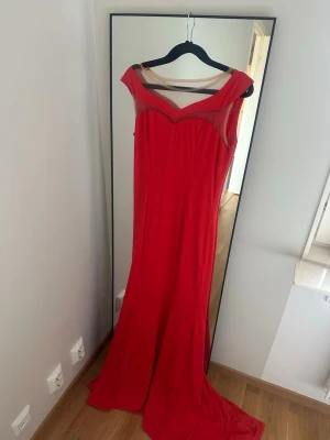 Röd långklänning med meshdetaljer - Elegant röd långklänning med transparenta meshdetaljer vid axlar och dekolletage. Klänningen har en figurnära passform och en vacker, svepande kjol. Perfekt för speciella tillfällen där du vill sticka ut.