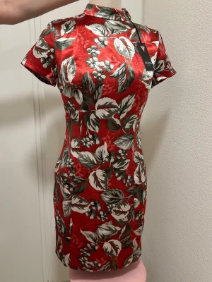 Röd blommig kortklänning Guess - Snygg röd kortklänning från Guess med blommigt mönster i vitt och grönt. Klänningen har hög krage, korta ärmar och figurnära passform. Tillverkad i ett glansigt material som ger en lyxig känsla. Perfekt för dig som vill sticka ut! Ny med prislapp. 