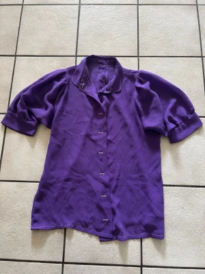 Lila blus med puffärm och krage storlek M/L - Snygg lila blus med puffiga korta ärmar och klassisk krage med dekorativ söm. Blusen har knäppning framtill och är tillverkad i ett lätt, flowy tyg som ger en cool vintagekänsla. Perfekt för dig som vill sticka ut med färg och stil.