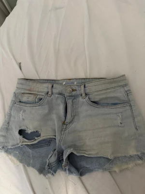 Ljusblå slitna jeansshorts lowrise - Säljer ett par ljusblå jeansshorts med råa kanter och flera slitningar för en edgy look. Shortsen har klassisk femficksdesign, dragkedja och knapp framtill. Perfekta för sommaren och ger en avslappnad vibe.