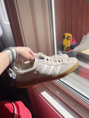 Adidas sambas - Säljer ett par klassiska Adidas sneakers i beige mocka med vita detaljer och de ikoniska tre ränderna på sidan. Skorna har en snygg gummisula och blå Adidas-logga på plösen. Perfekta för dig som gillar retrostil och streetwear.