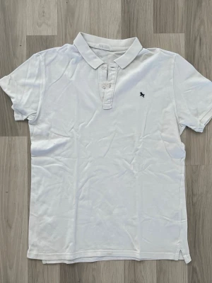 Vit piké från H&M - Klassisk vit pikétröja från H&M med liten broderad logga på bröstet. Tröjan har korta ärmar, krage och knappar framtill. Perfekt till jeans eller shorts för en clean och enkel stil.