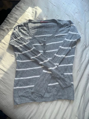 Gråvit randig ribbad långärmad topp - Snygg grå långärmad topp med vita horisontella ränder och ribbad struktur. Rund halsringning med knappdetaljer framtill ger en chill vibe. Perfekt till jeans eller kjol för en enkel men stilren look.