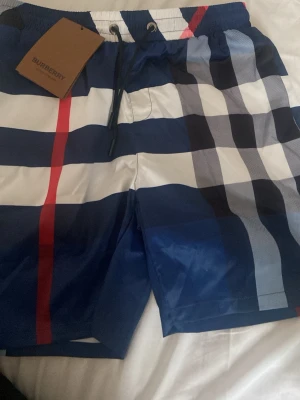 Burberry badbyxor blå-rutig - Snygga badbyxor från Burberry i blått med klassiskt rutmönster i vitt, svart och rött. Elastisk midja med dragsko och snabbtorkande material. Perfekta för stranden eller poolen med en modern och trendig look.