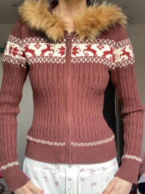 Brun stickad hoodie med fuskpäls - Mysig brun stickad hoodie med dragkedja och luva kantad med fluffig fuskpäls. Tröjan har vita och röda renmönster över bröstet och armarna samt kabelstickat mönster. Långa ärmar och figurnära passform. Perfekt för kalla dagar!