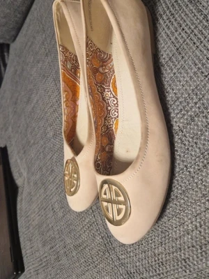 Beige ballerinaskor från Depeche - Snygga beige ballerinaskor från Depeche med rund tå och guldfärgad metallogo framtill. Skorna har ett dekorativt mönstrat innerfoder i orange och brunt samt knappdetalj bak. Tillverkade i ett mjukt material som ger en klassisk look.