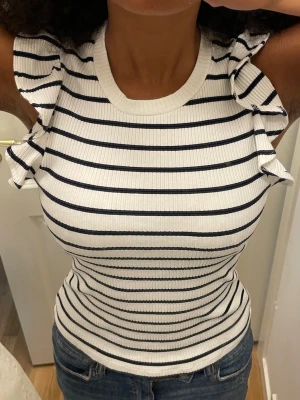 Randig ribbad topp med volangärmar - Säljer en vit ribbad topp med svarta horisontella ränder och volangdetaljer vid axlarna. Toppen är ärmlös och har en tight passform som framhäver figuren. Perfekt för dig som gillar en fräsch och trendig look. Har använt toppen ett fåtal gånger. Pris kan diskuteras