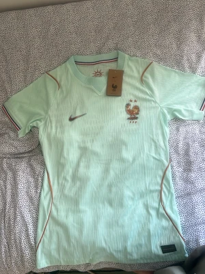 Frankrike Nike fotbollströja WM player version - Snygg mintgrön fotbollströja från Nike med Frankrikes landslagsmärke och två stjärnor på bröstet. Tröjan har korta ärmar, rund hals och tunna orange detaljer längs sidorna. Tillverkad i lätt och ventilerande material, perfekt för träning eller match.
