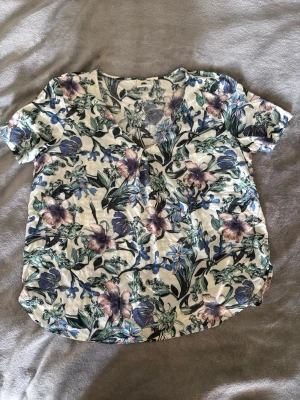 Blommig v-ringad topp - Kortärmad topp med v-ringning och blommigt mönster i grönt, blått och lila på vit botten. Tillverkad i mjukt bomullsmaterial och har en avslappnad passform. Perfekt för dig som gillar färg och mönster.