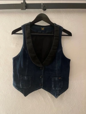Mörkblå jeansväst från Lee - Mörkblå jeansväst från Lee med svarta slag och två fickor framtill. Västen har knäppning med knappar och en klassisk, något dressad look. Perfekt för att lyfta din outfit med en cool detalj.