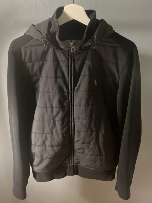Svart vindjacka från Polo Ralph Lauren - Snygg svart vindjacka från Polo Ralph Lauren med huva och dragkedja framtill. Jackan har quiltad front, släta ärmar och diskret logga på bröstet. Perfekt för blåsiga dagar och enkel att matcha med olika outfits.
