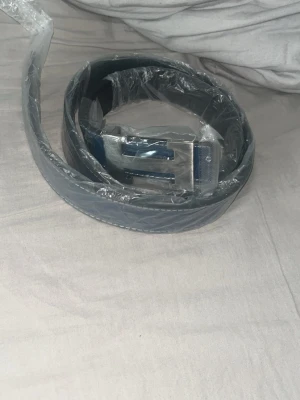 H Bälte  - Snyggt svart bälte med ett stilrent, silverfärgat metallspänne. Bältet har en klassisk design och passar perfekt till jeans eller kostymbyxor. Materialet ser ut att vara skinn eller syntet och spännet ger en modern touch.