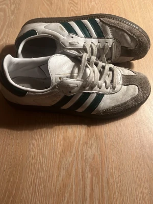 Adidas Samba vita och gröna sneakers - Säljer ett par klassiska Adidas Samba sneakers i vitt läder med gröna detaljer och grå mocka på tån och sidorna. Skorna har de ikoniska tre ränderna på sidan och Adidas-logga i guld på plösen. Perfekt för dig som gillar retrostil och sportiga vibes.