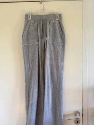 Gråa juicy couture byxor - Säljer ett par grå juicy byxor med raka ben och bred resår i midjan. Materialet är mjukt och skönt, perfekt för chill dagar. Passar dig som gillar en avslappnad stil. Dessa är i fint skick! Det är äkta. Lite missfärgat på detaljerna som hänger på snörena i midjan.