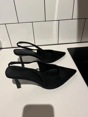 Svarta spetsiga slingback-pumps - Stilrena svarta pumps med spetsig tå och öppen häl med slingback-rem. Skorna har smal klack och är tillverkade i ett matt tygmaterial. Modellen är för lite bredare fötter därav lite bredare framtill. Jag har smal fot, men uppskattar dessa för att de är så sköna pga lite bredare framtill