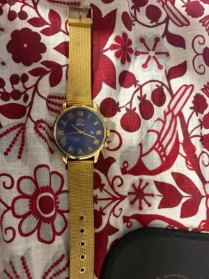 Guld klocka med blå urtavla - Stilren klocka från Curren med guldfärgat mesharmband och blå urtavla. Klockan har romerska siffror i guld, datumvisning och elegant rund boett. Perfekt accessoar för dig som vill sticka ut med en klassisk men modern look.