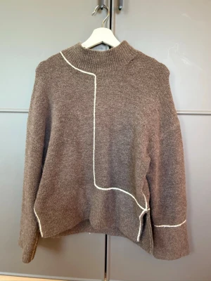 Brun stickad tröja med vit detalj - Säljer en brun stickad tröja med hög hals och unika vita kontrastsömmar som går över framsidan och ena ärmen. Tröjan har en avslappnad passform och långa ärmar. Perfekt för dig som gillar en modern och stilren look.🤎🤎🥥