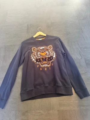 Kenzo sweatshirt med tigertryck - Cool svart sweatshirt från Kenzo med ikoniskt broderat tigerhuvud i orange, vitt och svart på bröstet samt texten 'KENZO PARIS'. Tröjan har rund halsringning och ribbade muddar. Perfekt för dig som vill sticka ut med en streetstyle-look.