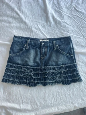 Jeanskjol med volanger - Säljer en kort jeanskjol med flera lager volanger nedtill. Kjolen har klassiska fickor fram och bak, bälteshällor och stängs med knapp och dragkedja. Perfekt för dig som gillar en edgy och lekfull stil.
