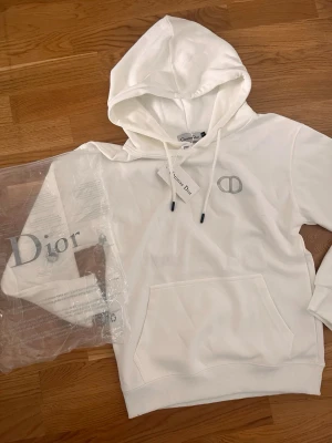 Vit hoodie från - Snygg vit hoodie från med broderad-logga på bröstet. Klassisk känguruficka framtill, justerbar huva med vita snören och ribbade muddar. Tillverkad i mjukt material som känns lyxigt och bekvämt. Perfekt för dig som gillar exklusiv streetwear.